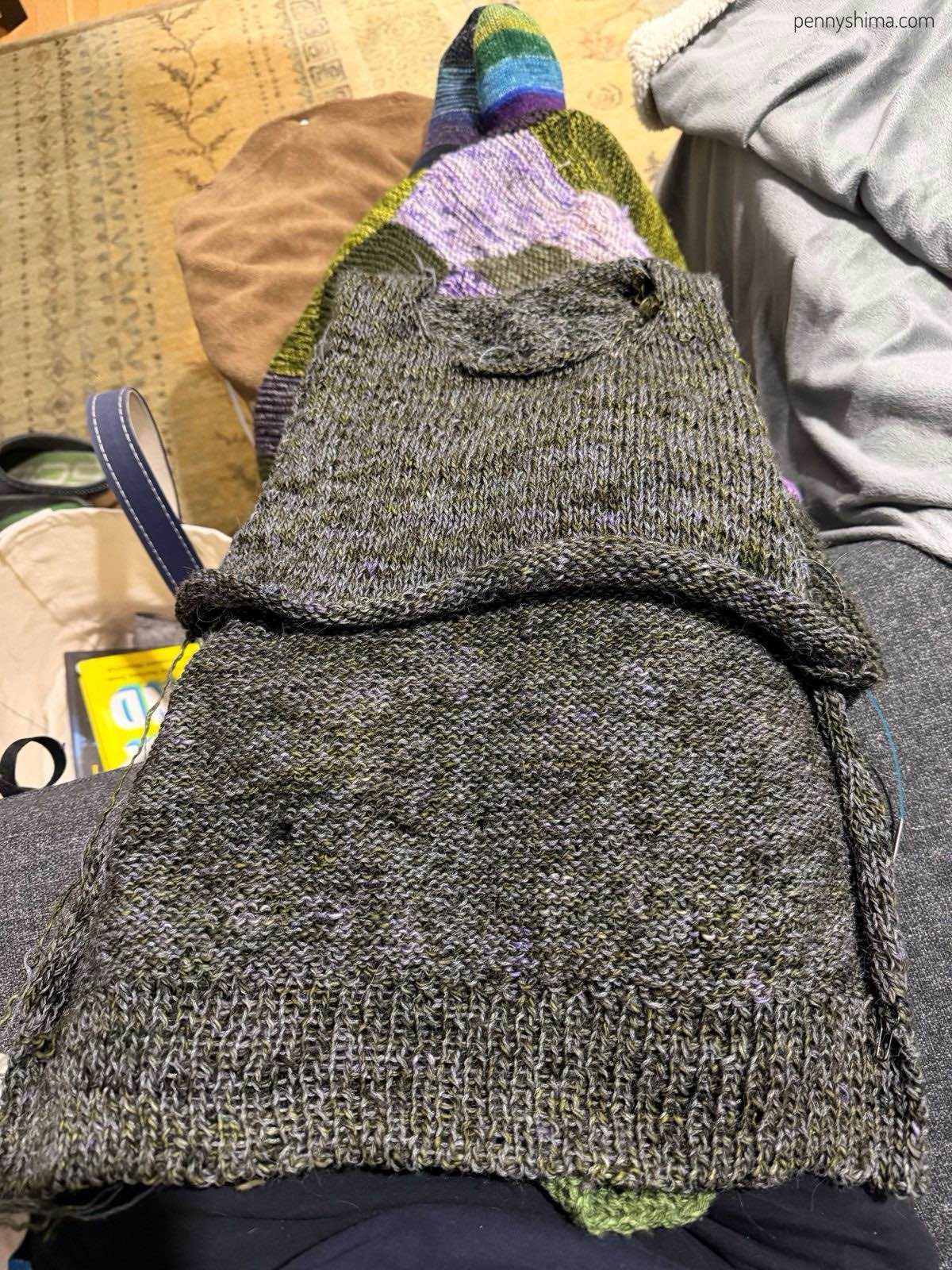 Lulu pullover (marled green with grey) after a few rows of armhole increases. It is folded and laying on the knitter’s outstretched legs