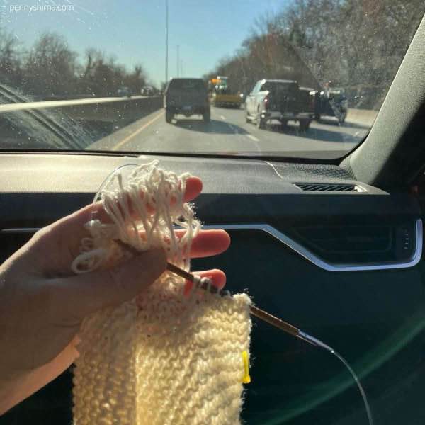 white knit selvedge being repaired held by a hand in the passenger seat of a car. a stitch marker is several rows below.