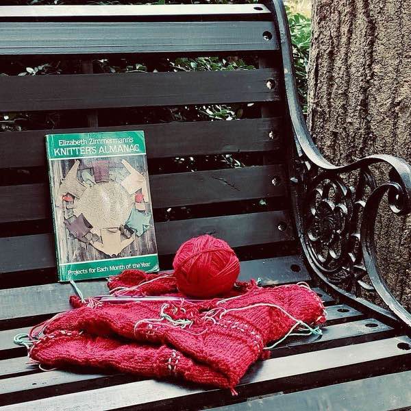 Red blanket in progress shown folded on a black wooden bench. A ball of yarn is on it, and copy of knitter’s almanac shown next to it