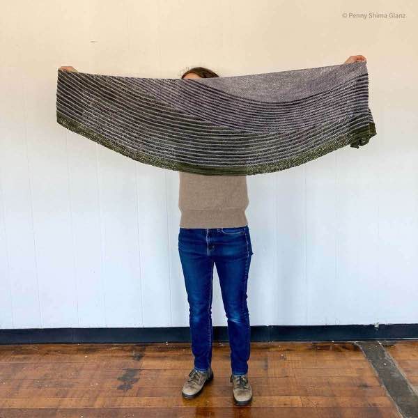 5 foot 2 inch person wearing jeans and a tan sweater holding up the grey and green striped shawl. They are standing in front of a white wall.