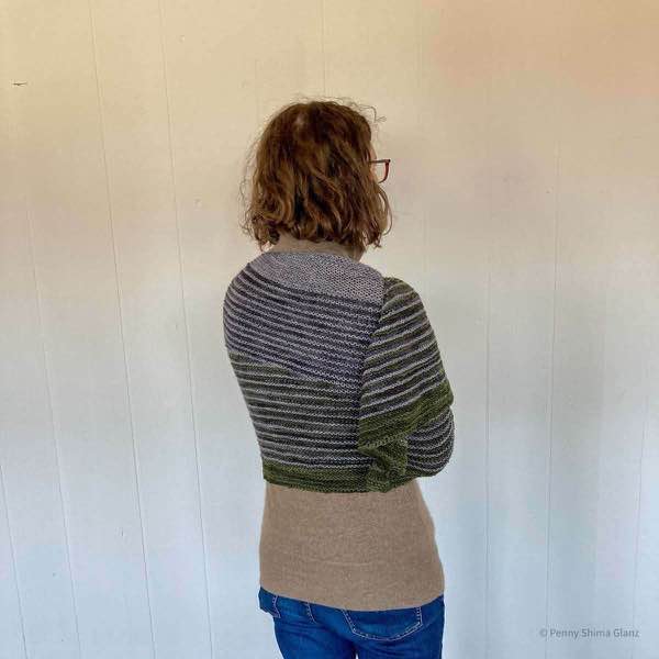 5 foot 2 inch person wearing jeans and a tan sweater Wearing the grey and green striped shawl. Their back is mostly to the camera and they are in front of a white wall.