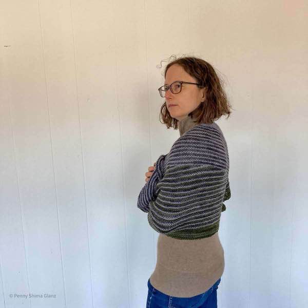 5 foot 2 inch person wearing jeans and a tan sweater with shawl wrapped tightly around the body. They are sideways to the camera and looking to the left.