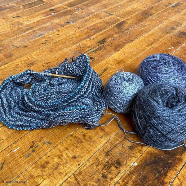 Grey and charcoal striped garter stitch knit shawl in progress shown on a distressed wooden floor next to the 3 cakes of yarn chosen for the project