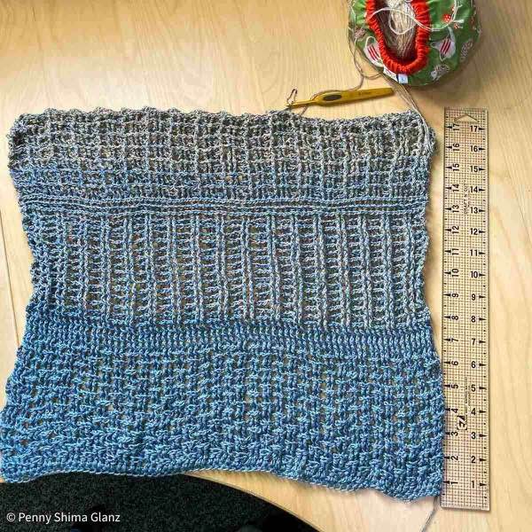Grey marled crochet wrap in progress , 18 inches / 45.75 cm shown on a light birch colored wooden desk next to a clear acrylic quilting ruler. The crochet hook is to in the active stitch (marked with a coil-less pin) and one of the cakes of yarn is shown above in a small bag. The project is in the third main pattern repeat, the 14th (of 15) rows.