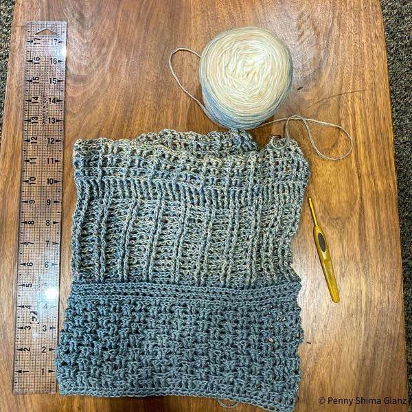 Grey marked crochet wrap in progress , 12 inches / 30.5 cm shown folded lengthwise on a wooden table next to a clear acrylic quilting ruler. The crochet hook is to the right and one of the cakes of yarn is shown above. The project is in the third main pattern repeat.