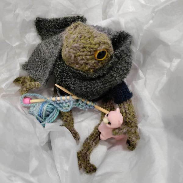frog wearing scarf, holding a pink alpaca (eraser) and working some light blue yarn into knitting.