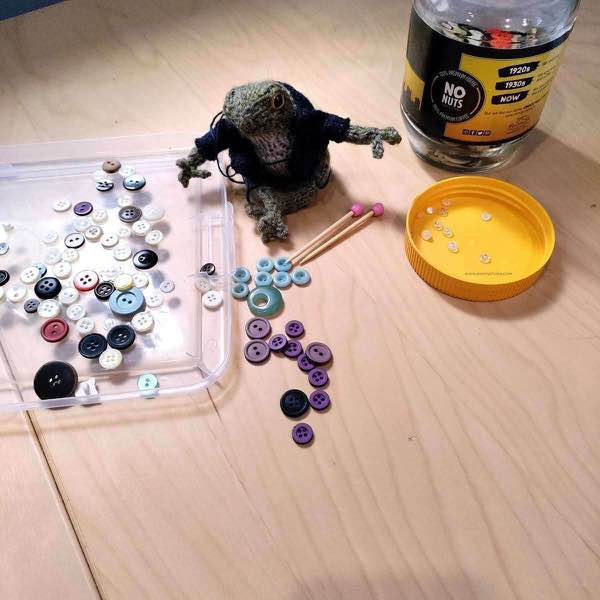 frog sitting on wooden desk next to buttons being sorted. some are in a old coffee jar with yellow lid, others on a plastic tray, some are on the desk.
