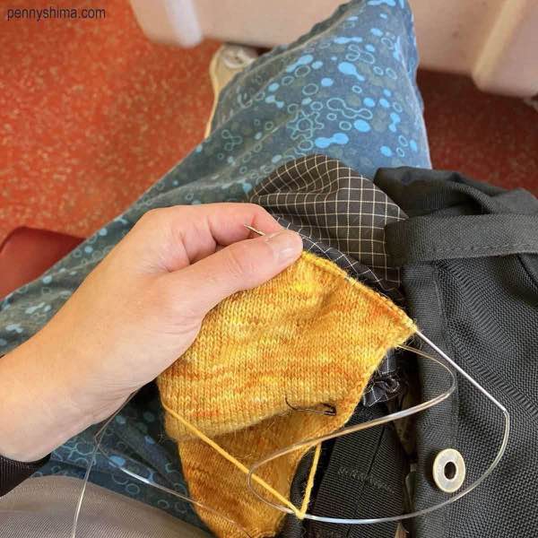 golden yarn being knitting into a sock, shown with the heel in progress. The knitter is sitting on a Metro North Train.