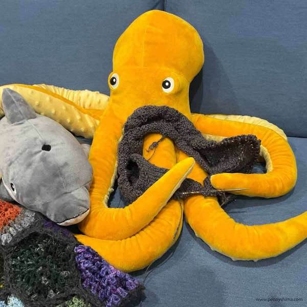 grey raglan cardigan in progress shown on a stuffed toy octopus that is a yellow-orange. there is a grey dolphin next to it. they are on a light blue couch.