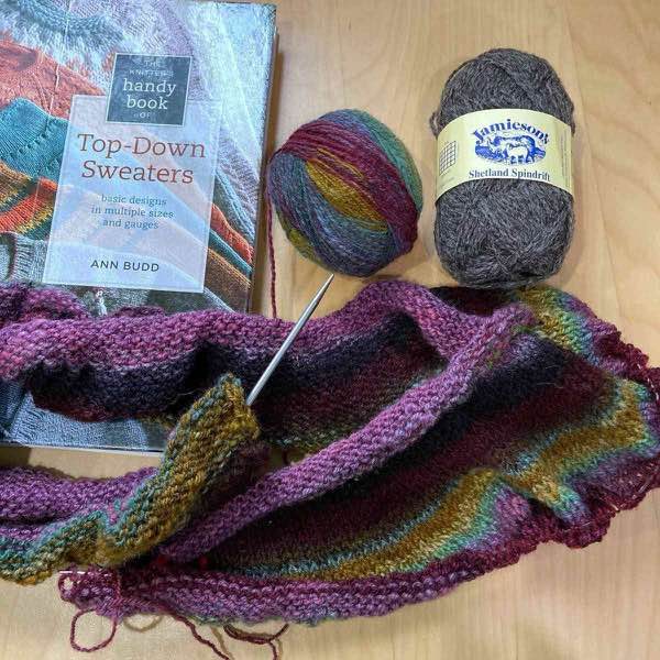 About 15cm of the knitted handspun yoke of sweater on needles laying on a wooden desk. The ball of handspun is next to the book, "The Knitter's Handy Book of Top-Down Sweaters" by Ann Budd. A skein of Jamieson's in Color 102 Shaela (a marled brown) is next to the handspun.