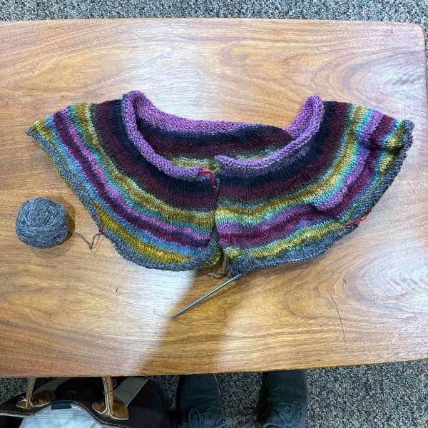 Muted rainbow handspun yoke, two rows past dividing for the sleeves. It is shown laid flat on a wooden table.