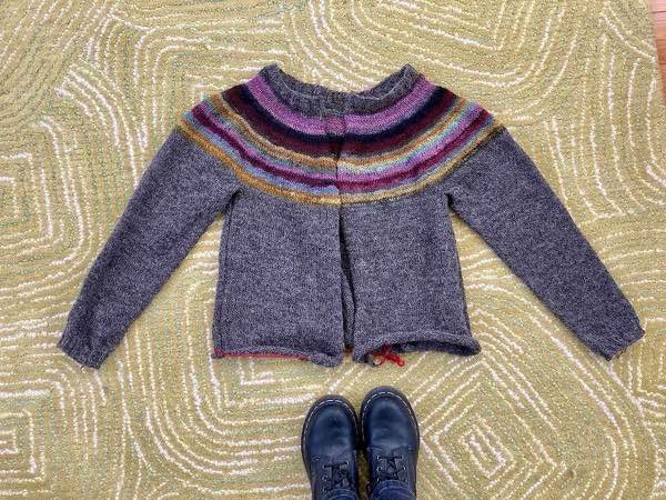 Muted Rainbow handspun yoke cardigan with sleeves and neck band complete. Shown laid flat on a wool rug of green/white outline rectangle motifs and the knitter's black doc martens.