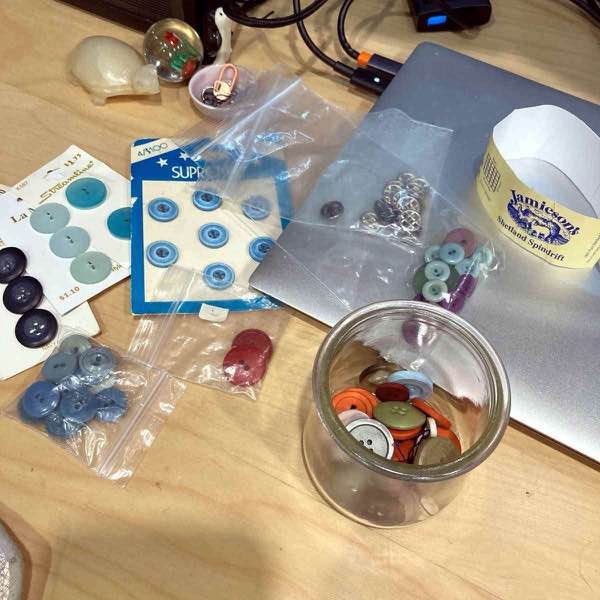 various buttons in a rainbow of colors and sizes shown on cards and in small plastic bags and a clear glass jar strewn on a wooden desk. a ball band for jamieson's shetland spindrift is also visible.