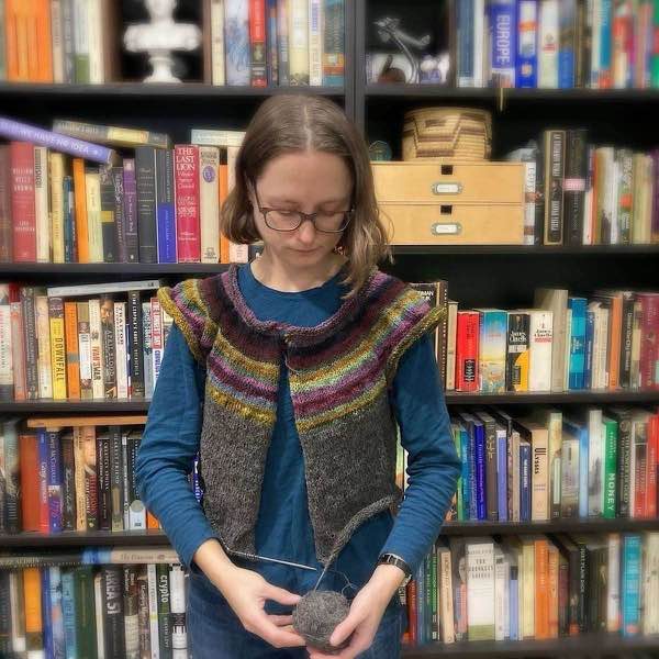 Muted rainbow handspun yoke. Shown modeled with 20cm of body knit. Model is wearing a blue T-shirt and is standing in front of bookshelves. The model is holding a ball of yarn and the knitting needles are visible. The background is slightly blurred.