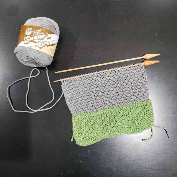 A grey and green knit washcloth in progress on a black surface