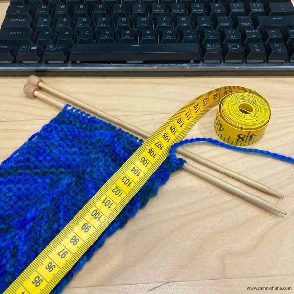 small section of the blue stripe of blanket in progress with a yellow tape measure showing 107 cm knit. The knitting is on a light wood coloured desk and a black keyboard is visible.