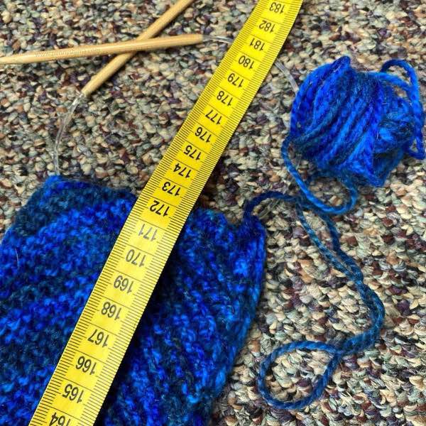 yellow tape measure showing length at 172cm. the NE blanket is in a blue yarn and is on a short bamboo circular needle. It is on a carpet of browns and neutral colours