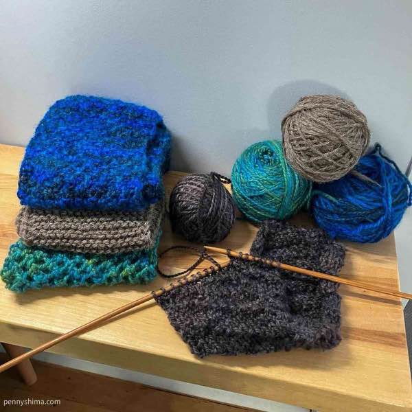 pile of 3 complete blanket stripes (bottom to top: aqua, light brown, royal blue) on a wooden surface next to the balls of yarn. In front a dark brown stripes is shown on the needles.