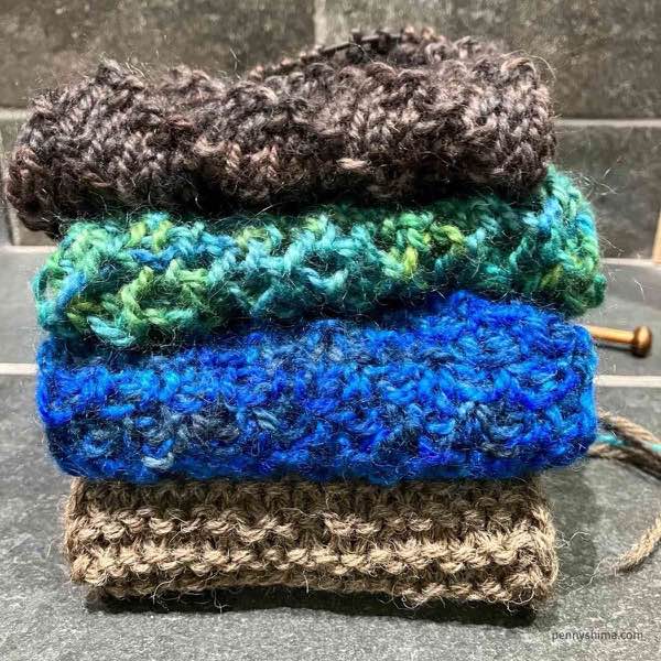 Stack of knitted stripes on a grey tile surface. Bottom to top: light brown garter stitch, blue moss stitch, aqua lace, brown purl diagonal