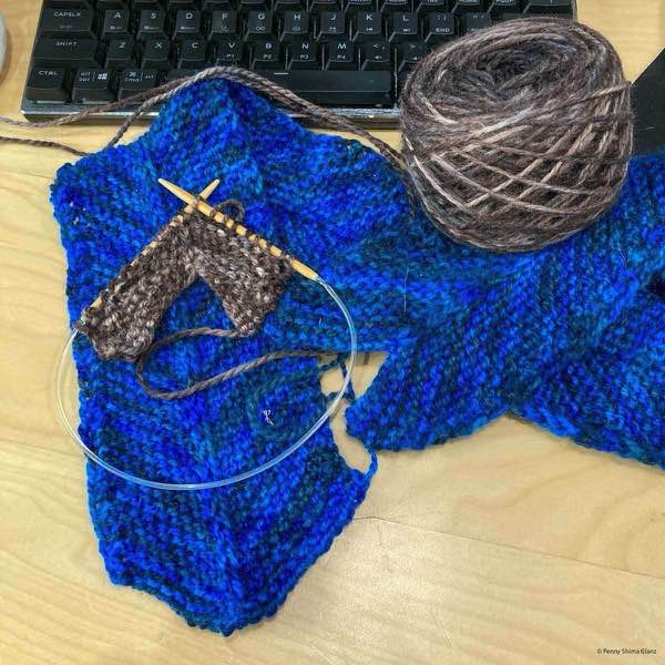 second column of library north eastrly banket in progress shown on a birch desk with the first stripe shown folded over itself. The brown yarn is caked on top and is shown laying on top. a computer keyboard is in the background.