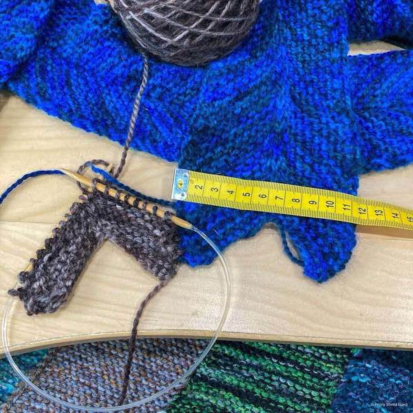 NE library blanket in progress on a birch wooden desk. The second column in a dark brown is on it's 5th ridge. A yellow tape measure is laid across the first column, it shows about 11cm in width.