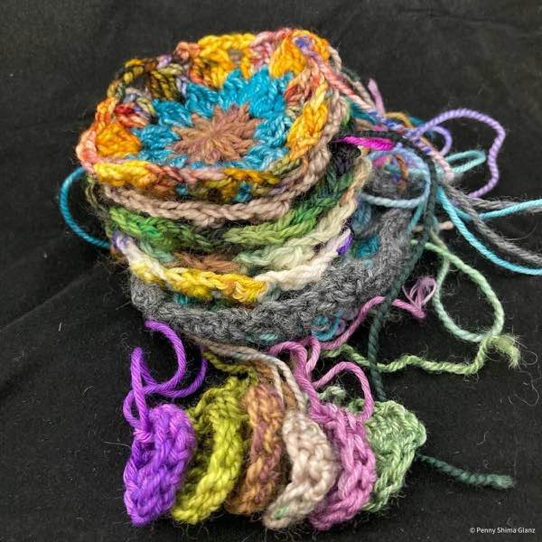 stack of motifs for the bauble crochet blanket on a black background. in the foreground are six centers. theyu are a range of colors but only the top motif is fully visible, it has a brown center, teal second round, and a multi of gold-blues-reds for the third round. the centers are purple: lime green, brown, mostly ecru, rose pink, and a light green. the yarn ends are not woven in.
