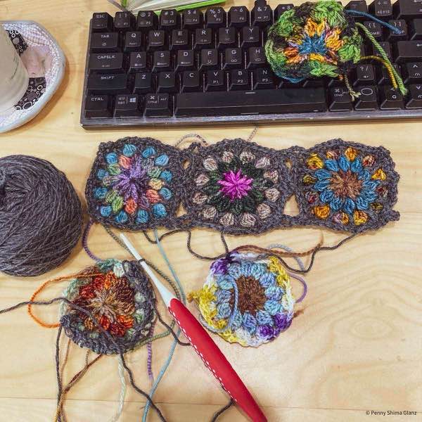 Motifs on a light color wooden desk. The first three motifs are joined with grey yarn. The fourth is being worked and shown with the crochet hook in progress.