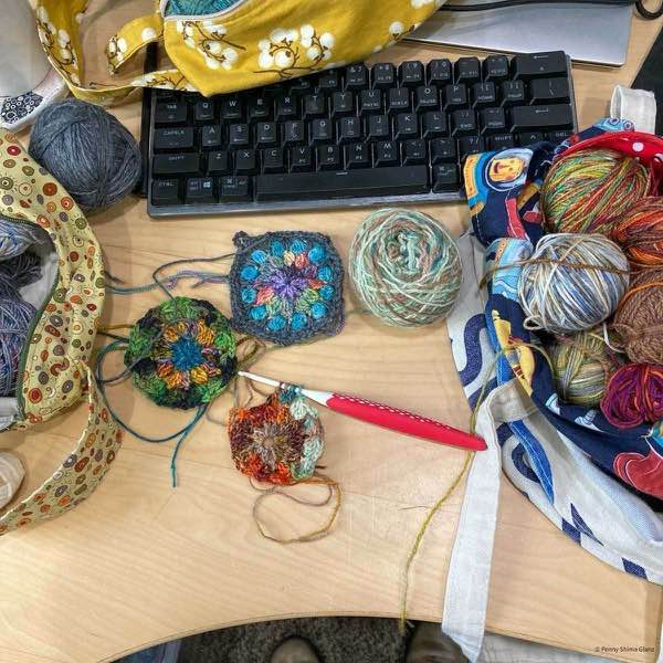 several crochet motifs and balls of yarn messy piles on a birch desk with a computer keyboard