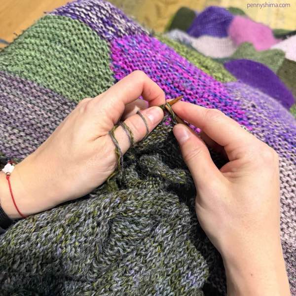 hands holding knitted fabric, the knitter is purling. it's a grey-green marled fabric (design is lulu slipover by petite knit)