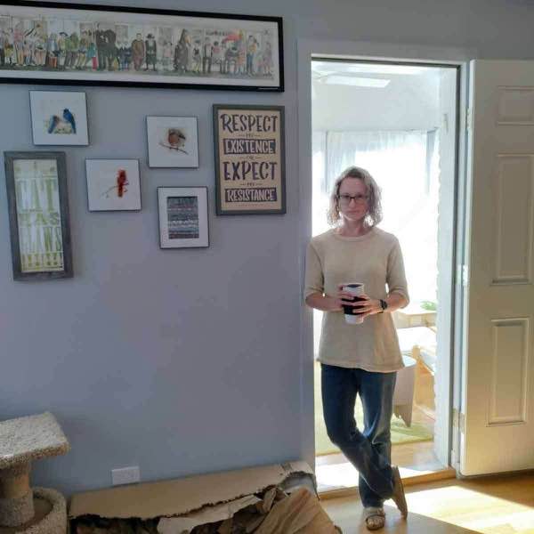 completed tee modelled, front. psg next to artwork hanging on a light green wall. the model is back lit and is holding a travel coffee mug (full of coffee made at home)