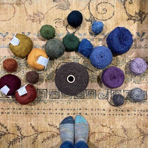 a rainbow of wool yarn in balls and cakes shown around a cone of dark brown and a light gold and green patterned rug. The photographers feet are visible and they are wearing hand knit socks.