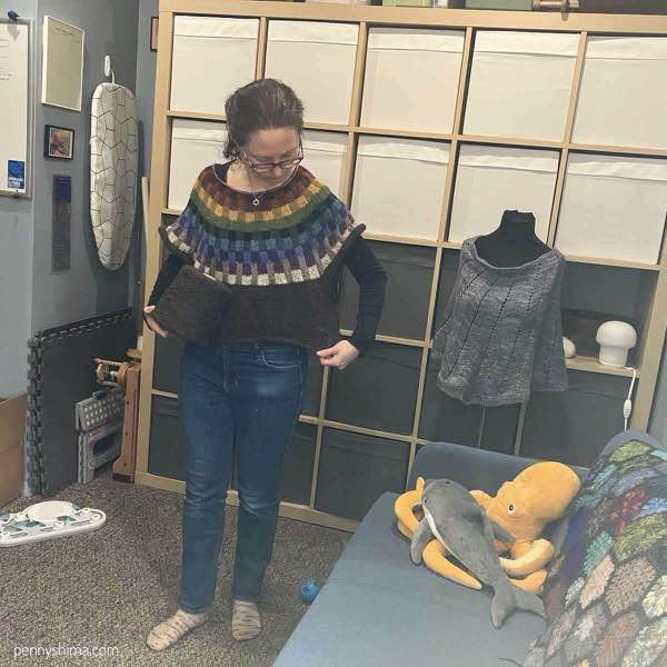 Paul Klee sweater tried on while on the needles. The model is in their studio office space and is holding the cone of yarn and stretching some of the body from the left.