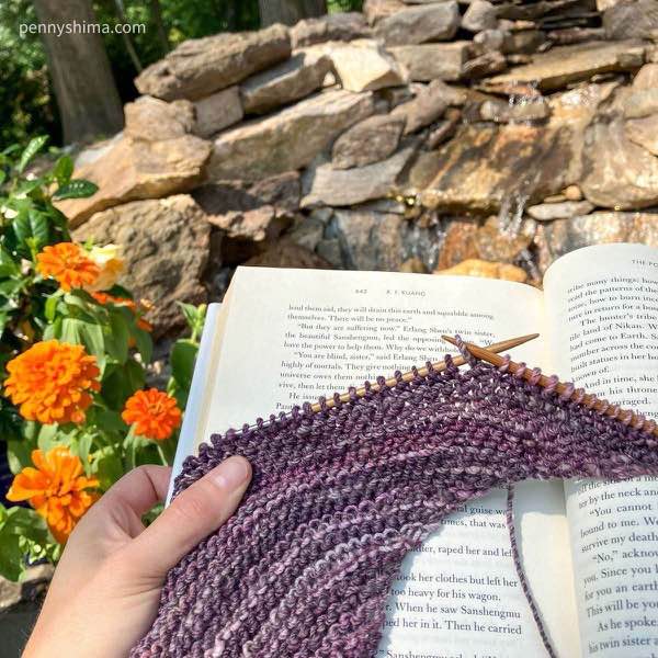 a hand knit shawl in progress out of purple handspun being held on “the poppy wars” by k f kuang p 442 (no significance to this page other than that’s where I am) next to orange flowers and in front of a small water feature