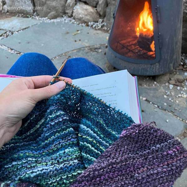 a hand knit shawl in progress out of handspun being held on an open book in front of a chimenea with a small fire
