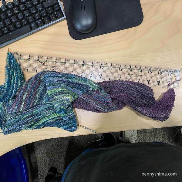 a hand knit shawl in progress out of purple, blues, and blue-green handspun on a wooden desk next to a keyboard. a strand of green and blue yarn is held in place by a clear quilting ruler. the yarn measures 18in (45.75 cm) in length.