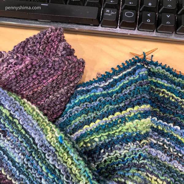 a hand knit shawl in progress out of purple, blues, and blue-green handspun on a wooden desk next to a keyboard.