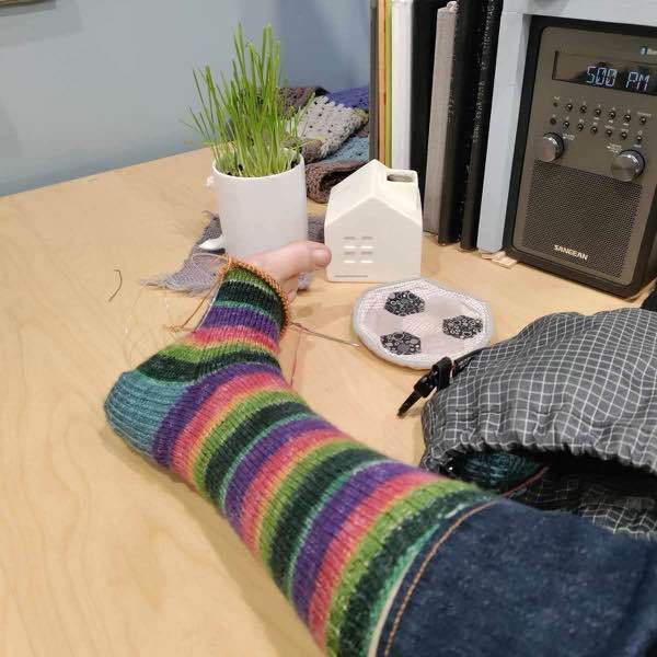 Rainbow sock on needles and a foot that is resting on a desk. there's a few rounds to go before the toe.