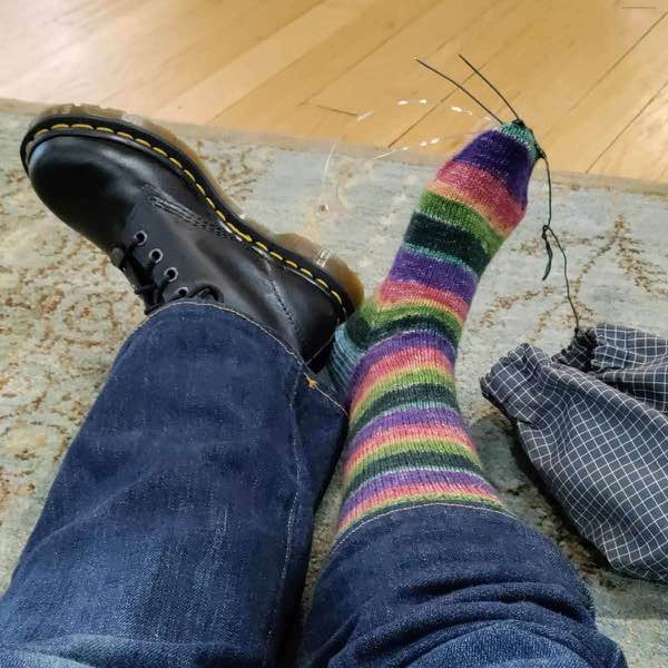 Rainbow sock on needles and a right foot that is resting on the carpet. The sock toe is finished and needs to be grafted.
