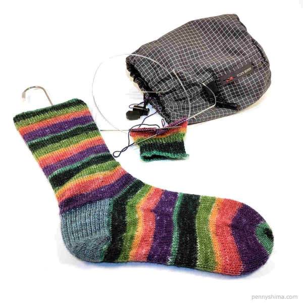 one completed rainbow sock on wire sock blocker, second sock started and knit about an inch past cuff. grey tom bihn project bag shown. all on plain white background.
