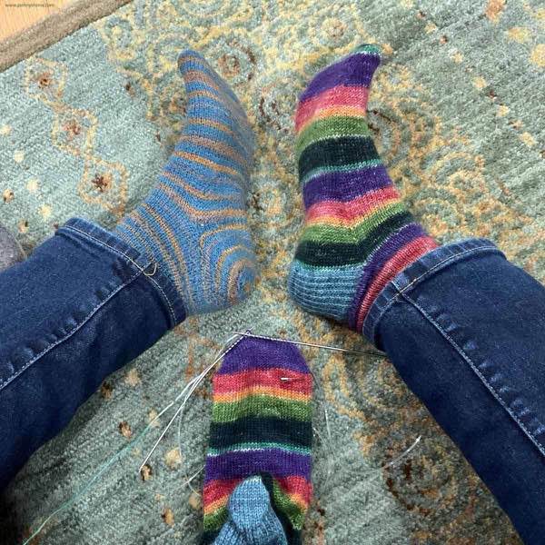 Two different socks worn modelled while the toe of the second Rainbow sock is knitted. the person is sitting on a light brown and green rug.