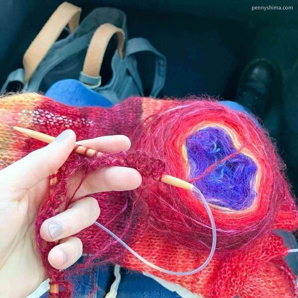 artyarns silk mohair ombre 2ply cardigan in progress. the color being worked is red, the yarn cake is visible with red transitioning to gold and then apurple.