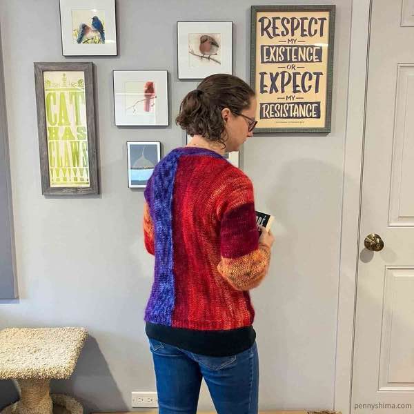 knitter wearing the sandstone striations cardi in front of a light grey wall with artwork. back right side.