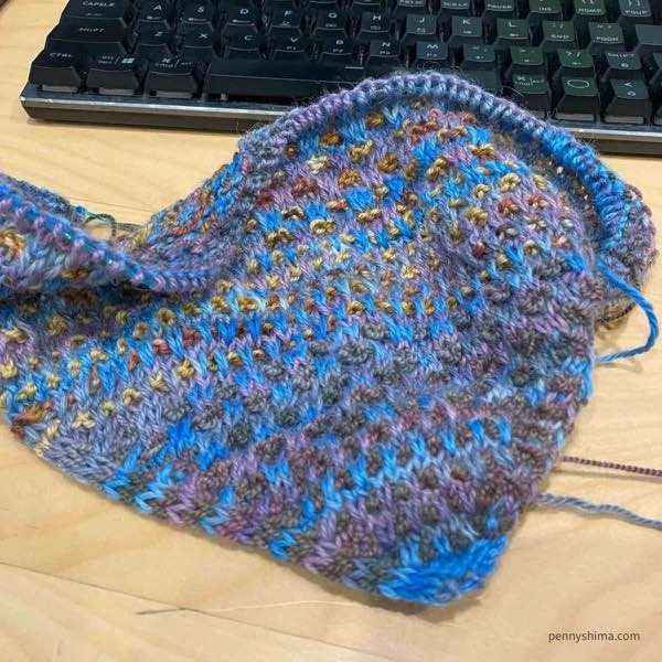 Shift cowl in progress (Section 3) shown on a wooden desk in front of a keyboard. Color A is blues and purples. Color B is muted marroon and browns and greens, Color C is a golden straw with rose and browns