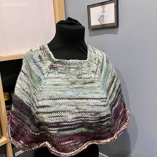 Silky twist poncho in Sierra blooms on a black fabric covered mannequin/dress form. The cast on is light blues/greens and is it segues to purples and deeper greens with hints of yellow.