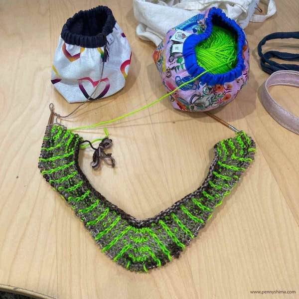 KAL top, front progress laid out on a wooden desk with two balls of yarn in fabric bags shown nearby
