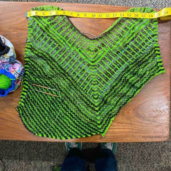 KAL top laid flat on a wooden table with a measuring tape across the shoulders to show width. One corner is squared off and the rest is in progress.