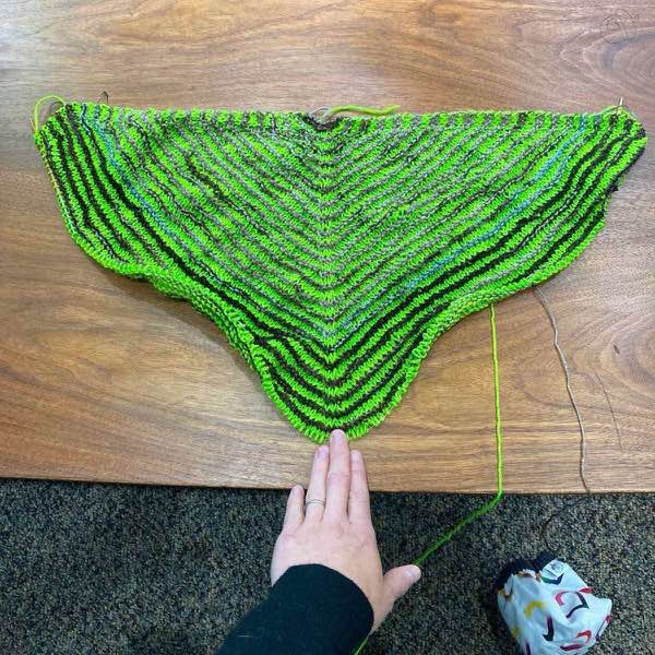 Green and brown back of KAL top shown laid flat on a wooden table. About 25 Rows complete. The knitter's hand is holding the center point.