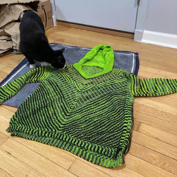Tuxedo cat sniffing the just blocked KAL sweater laid flat on a blue towel and wooden floor