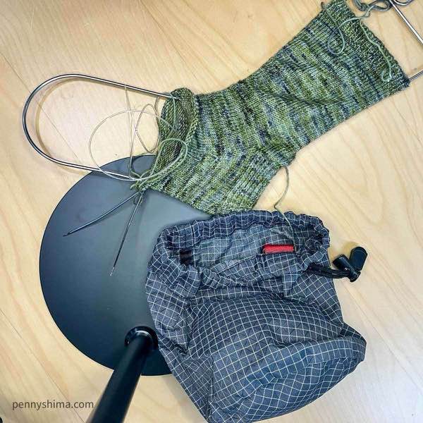 Spring green varigated socks in progress, heel is turned and gussets completed. sock is on a sock blocker and shown flat on a light wooden desk. a grey tom bihn stuff sock is next to it.