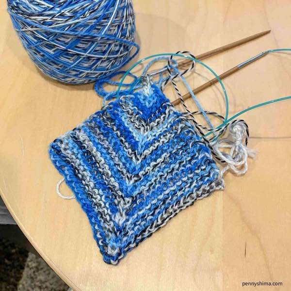 Miter square in blue zebra stripe yarn shown on needles and on a light color wooden surface